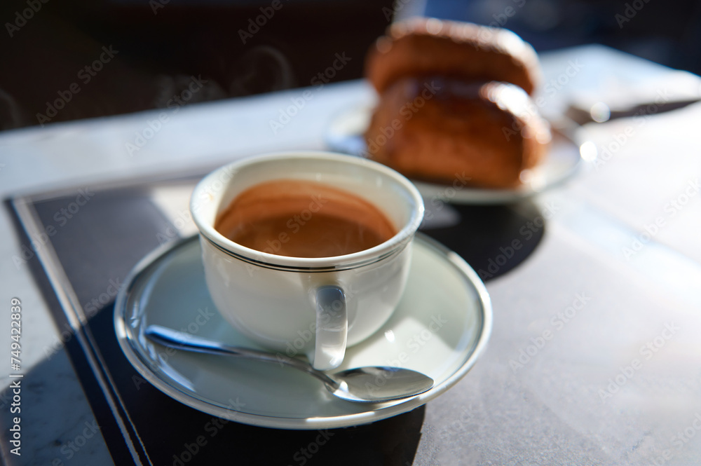 Elegant white ceramic cup of freshly brewed espresso coffee with foam on saucer. Blurred crispy croissants on background Elegant white ceramic cup of freshly brewed espresso coffee with foam on saucer. Blurred crispy croissants on background