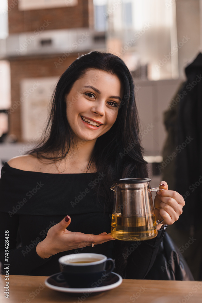 Brunette woman sitting at table drinking tea in restaurant. Teen woman wear black long sleeves top hold teapot with green tea, cups in cafe. Happy girl relaxing and rest with friends.