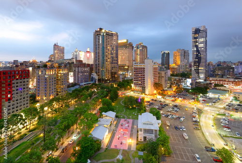 Night skyline of Downtown Taichung, a vibrant metropolis in central Taiwan, with modern high-rise buildings standing by a wooded green boulevard & dazzling city lights glowing under blue twilight sky 