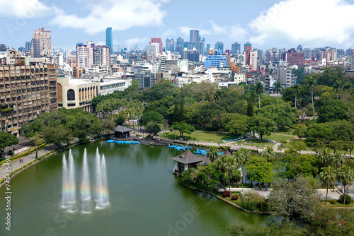 Aerial scenery of Downtown Taichung, a vibrant metropolis in central Taiwan, with modern skyscrapers in background & a rainbow fountain in a pond surrounded by green forests in a park under sunny sky © AaronPlayStation