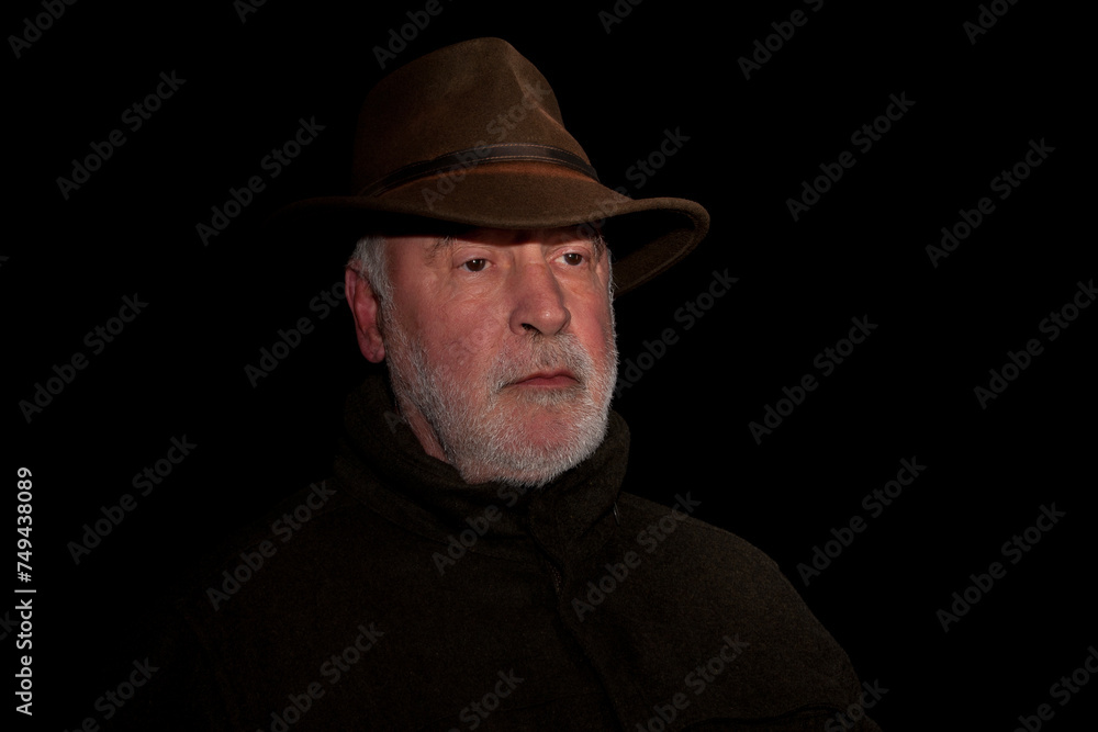 Portrait of a thoughtful looking old hunter with a beard who exudes calm and his face marked by experience, against a black background.