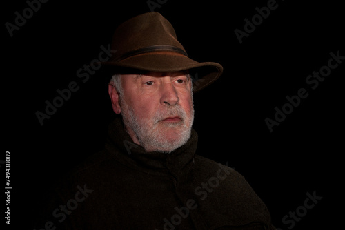 Portrait of a thoughtful looking old hunter with a beard who exudes calm and his face marked by experience, against a black background.