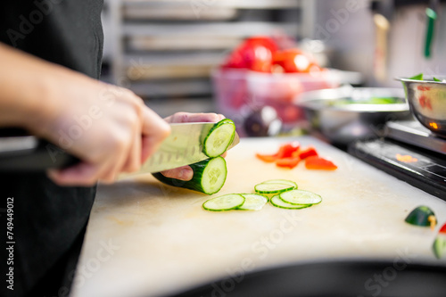 A chef deftly slices fresh cucumber on a cutting board in a professional kitchen. Chopped vegetables surround the scene, and stainless steel surfaces gleam in the background.