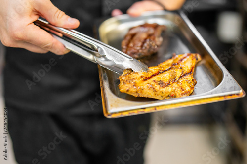 Close-up of a chef holding a tray with grilled chicken and steak using tongs. The chef, dressed in black, serves the deliciously grilled food. photo