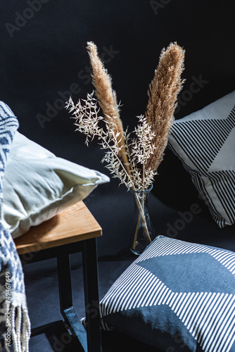 interior and home decor concept - close up of pillows and blanket on bench and dry plants in vase on floor in dark room