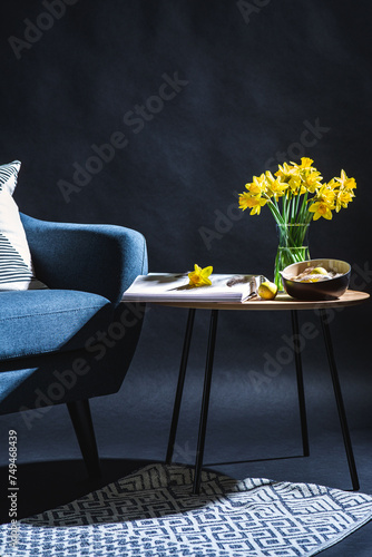 interior, holidays and home decor concept - modern blue chair with pillow, easter eggs hanging in bowl and daffodil flowers on table in dark room