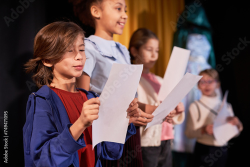 Side view portrait of young boy rehearsing lines standing on stage in school theater in row with children actors