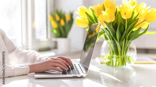 Female hands typing on a laptop, close-up. A woman works, sitting at a table at home, in the office. There is a bouquet of yellow tulips on the table. Women's Day, Mother's Day © Irina