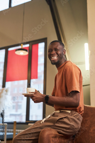 Vertical portrait of smiling Black man with cup of coffee looking at camera cheerfully in coffee shop