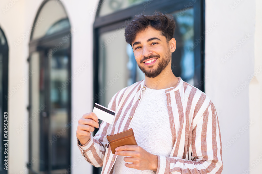 Handsome Arab man at outdoors holding wallet and credit card with happy expression Handsome Arab man at outdoors holding wallet and credit card with happy expression