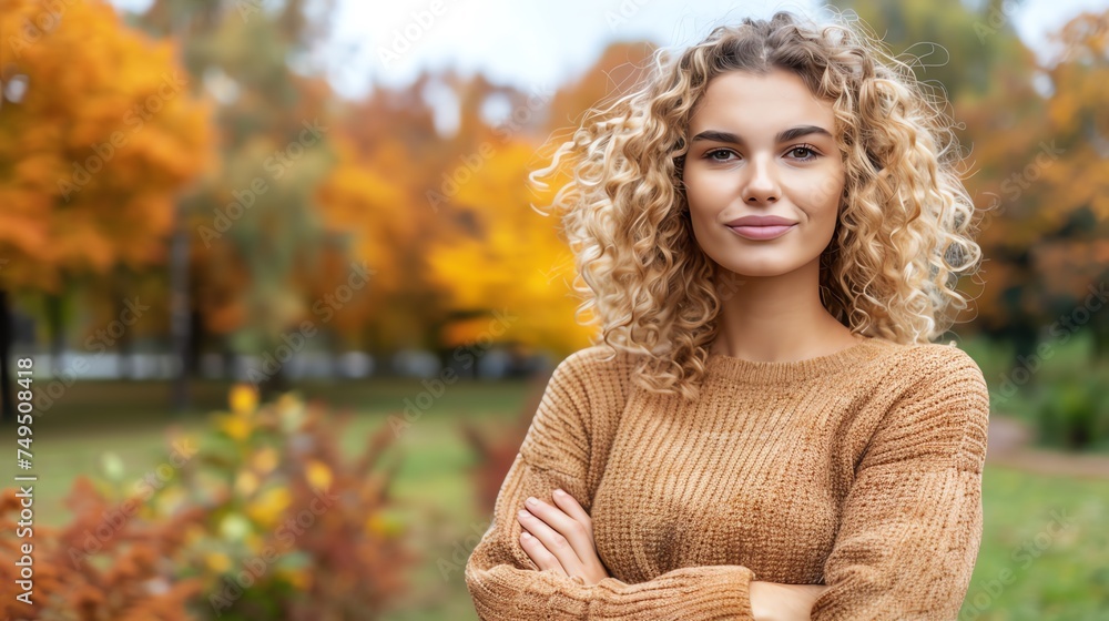 A young woman with curly blonde hair exudes warmth and confidence against an autumnal park backdrop. Her cozy, knitted sweater blends seamlessly with the golden hues of the fall foliage.