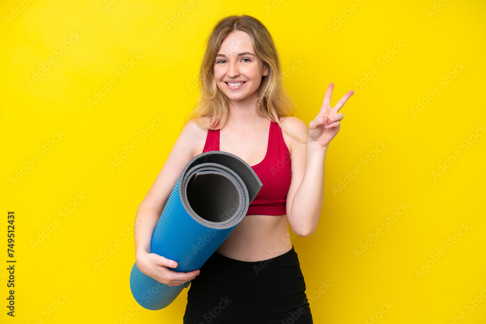 Young sport caucasian woman going to yoga classes while holding a mat isolated on yellow background smiling and showing victory sign Young sport caucasian woman going to yoga classes while holding a mat isolated on yellow background smiling and showing victory sign