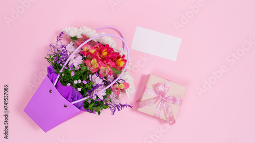 Bouquet of fresh flowers in purple bag along with white card and gift box on pink background.