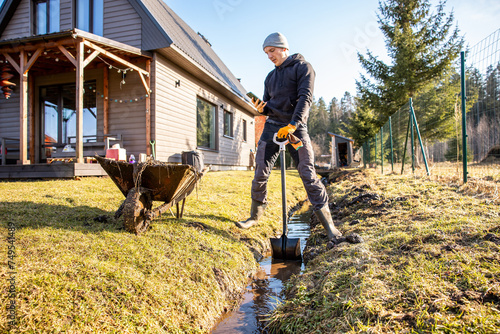 Man pausing from outdoor work to check his phone while kneeling by a newly dug drainage ditch in a lush backyard setting. photo