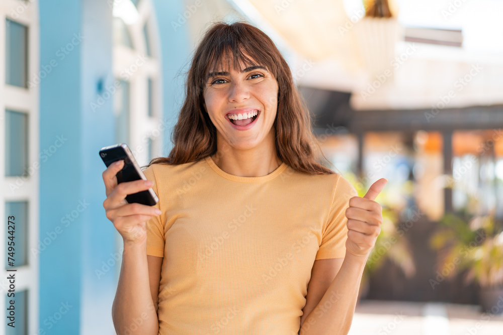 Young woman at outdoors using mobile phone while doing thumbs up Young woman at outdoors using mobile phone while doing thumbs up