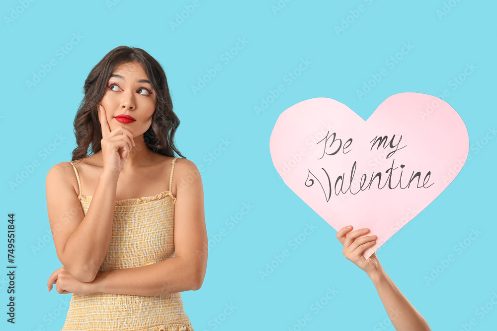 Beautiful young thoughtful Asian woman and hand with paper heart on blue background. Valentine's Day celebration Beautiful young thoughtful Asian woman and hand with paper heart on blue background. Valentine's Day celebration