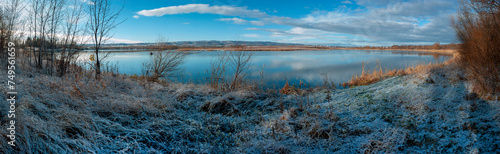 Panorama of the sunrise over the winter river. The sun rises over the lake. Autumn frost. The frozen river is covered with thin ice, the grass glistens with frost.