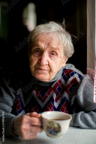 An elderly woman sits at a table with a cup in her hand.