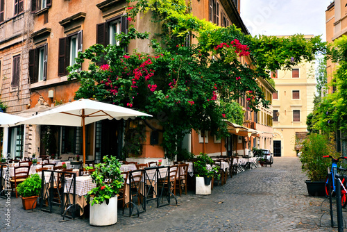 Fototapeta Naklejka Na Ścianę i Meble -  Beautiful ancient street in Rome lined with leafy vines, flowers and restaurant tables, Italy