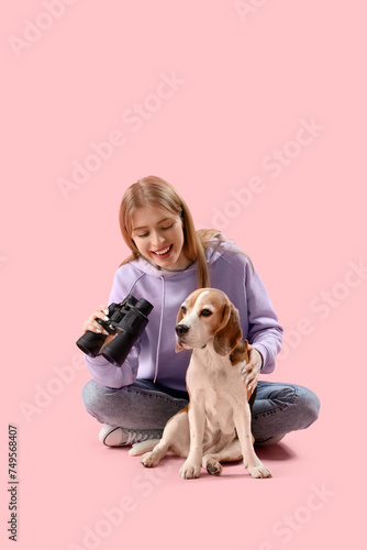 Young woman with binoculars and Beagle dog sitting on pink background