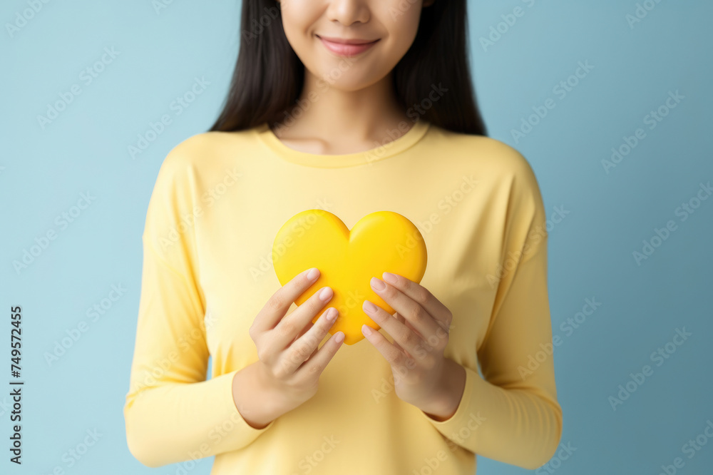 Young woman in yellow holding a heart-shaped object, symbolizing love and affection on a blue backdrop. Young woman in yellow holding a heart-shaped object, symbolizing love and affection on a blue backdrop.