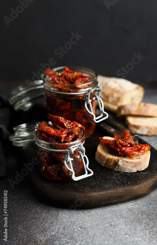 Sun-dried tomatoes in a glass jar. Tomatoes with spices. Dark background. Summer dish. Italian cuisine. Sun-dried tomatoes on white bread.