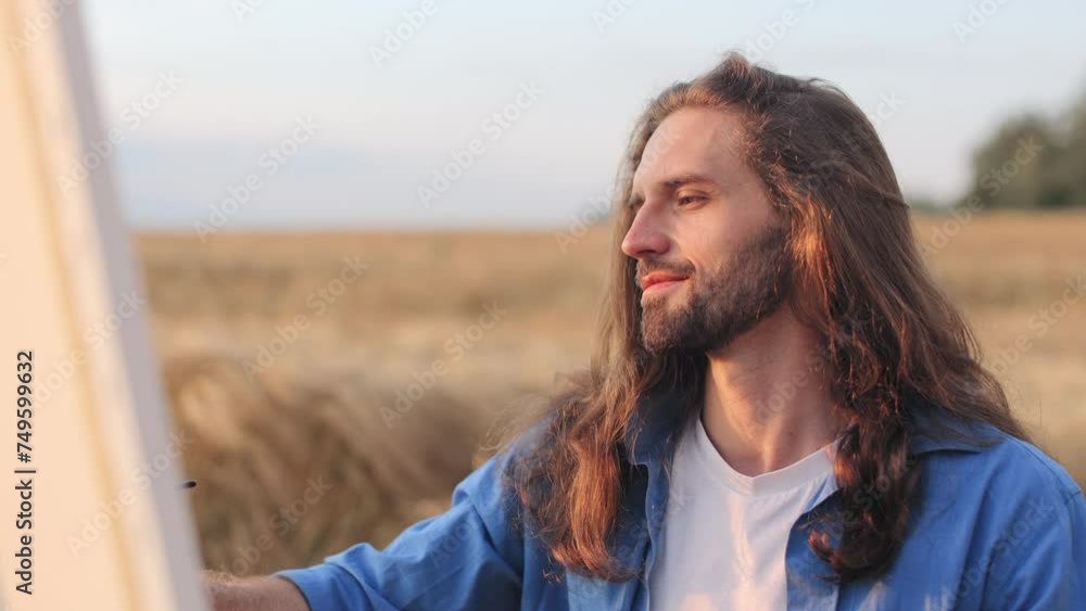 Close up of smiling male artist with long hair sitting by easel and painting on canvas while enjoying sunset light. Talented painter working and feeling satisfied with process surrounded by nature. Close up of smiling male artist with long hair sitting by easel and painting on canvas while enjoying sunset light. Talented painter working and feeling satisfied with process surrounded by nature.