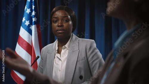 Medium shot of young African American female politician having confrontation with anonymous opponent at debate, in front of USA flag, blaming and suggesting solutions to high inflation crisis photo