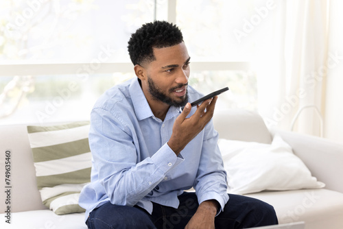 Serious young Black business man holding smartphone at mouth, dictating voice command at speaker, speaking audio message at mobile phone, activating virtual assistance app