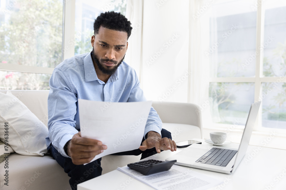 Serious young African American man calculating expenses, income at home, typing on calculator, using laptop, reading legal financial document, doing accounting paperwork