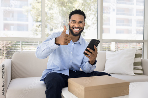 Cheerful satisfied young African client man making like hand gesture, showing thumb up at camera, smiling for portrait, using transportation online service over logistic cardboard box photo