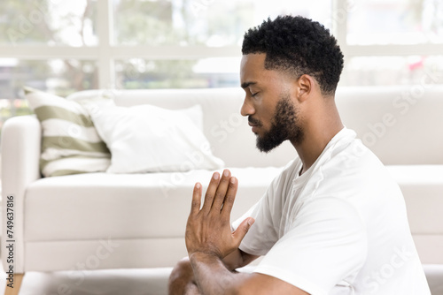 Focused calm African American guy meditating at home, practicing pranayama, breath work, keeping Namaste greeting hands gesture, sitting on floor with closed eyes. Side view