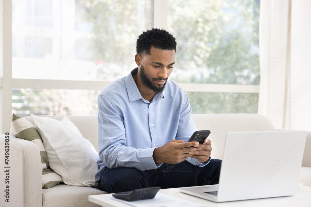 Focused busy young African man doing accounting job at home, using mobile phone at laptop and calculator, typing, counting budget with financial application, checking online money balance Focused busy young African man doing accounting job at home, using mobile phone at laptop and calculator, typing, counting budget with financial application, checking online money balance