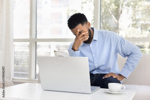 Exhausted frustrated African remote employee man sitting at laptop computer on home couch, touching head, covering face, getting bad news, online app problems, technologies failure