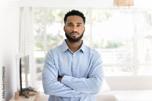 Serious confident handsome African freelance business man in office shirt standing indoors with hands folded at chest, looking at camera. Young male entrepreneur, homeowner home portrait photo