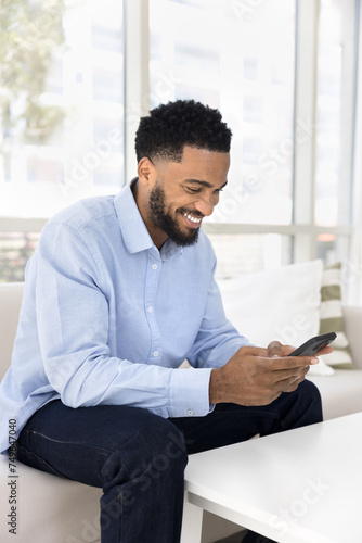 Cheerful young African man chatting on mobile phone, typing message on digital gadget, sitting on home couch, using Internet technology, online business communication, smiling, laughing