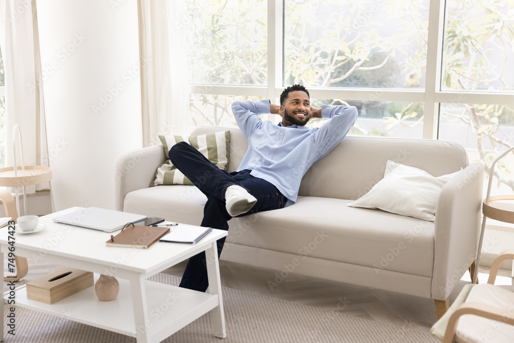 Cheerful young black freelance employee man stretching body on white comfortable home couch in cozy interior, smiling, looking away, enjoying leisure, relaxation, taking work break, pause Cheerful young black freelance employee man stretching body on white comfortable home couch in cozy interior, smiling, looking away, enjoying leisure, relaxation, taking work break, pause