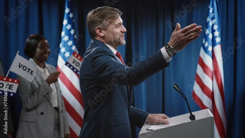 Medium side shot of middle-aged Caucasian male US president or senator walking onto stage towards podium, greeting audience and delivering energetic keynote address while campaigning for reelection photo