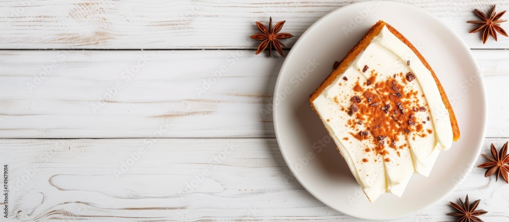 A slice of pumpkin cake with cream frosting sits on a white plate on a white wooden table, viewed from above.