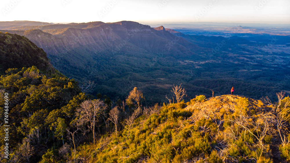 aerial view of hiker girl standing at the top of mountain, enjoying sunset over unique, folded mountains in south east queensland, australia; main range national park near brisbane, bare rock lookout	