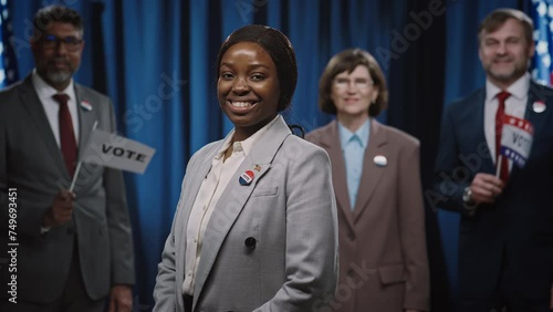 Medium portrait shot of cheerful African American female politician running for presidency, Senate or Congress, posing in front of USA flag, adjusting suit jacket, smiling, with diverse campaign staff photo