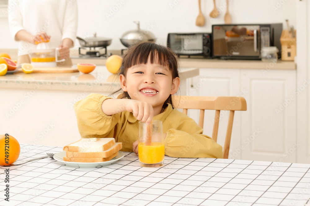 Asian woman making orange juice in kitchen, girl drinking juice and eating bread Asian woman making orange juice in kitchen, girl drinking juice and eating bread
