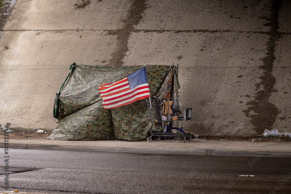 Φωτογραφία An American flag at a homeless tent made of camouflage tarp at a road underpass