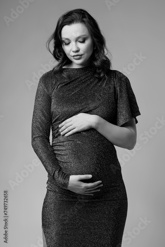 Black and white portrait of young pregnant female in grey sequin dress with hands near pregnant belly.