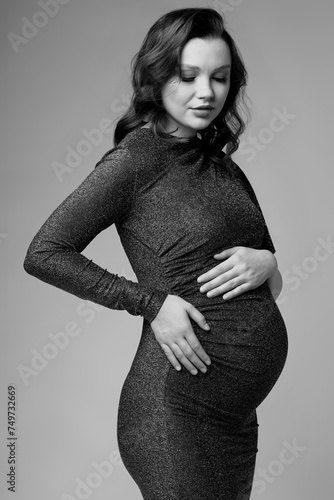 Black and white portrait of young pregnant female in grey sequin dress with hands near pregnant belly.