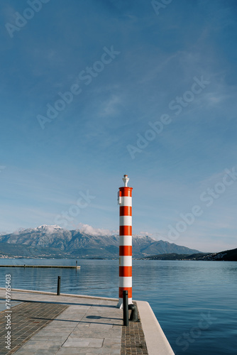 Small striped red and white lighthouse on a pier by the sea against the backdrop of mountains © Nadtochiy
