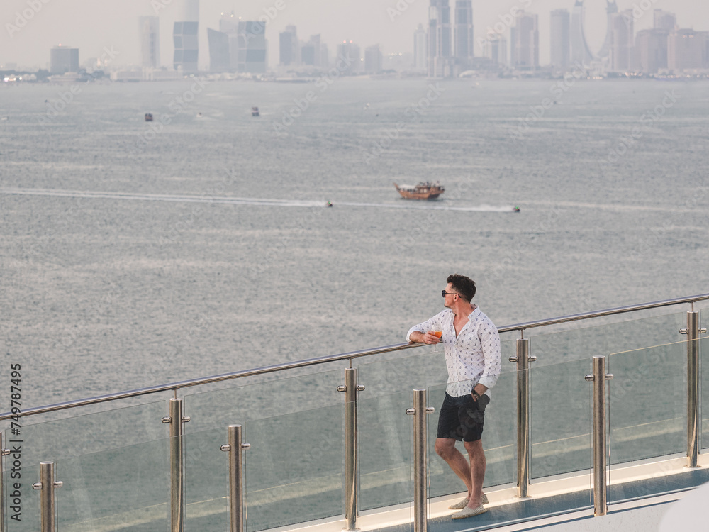 Stylish man with a glass of drink standing on the empty deck of a cruise liner against the backdrop of sea waves. Sunny, clear day. Close-up, outdoor. Vacation and travel concept