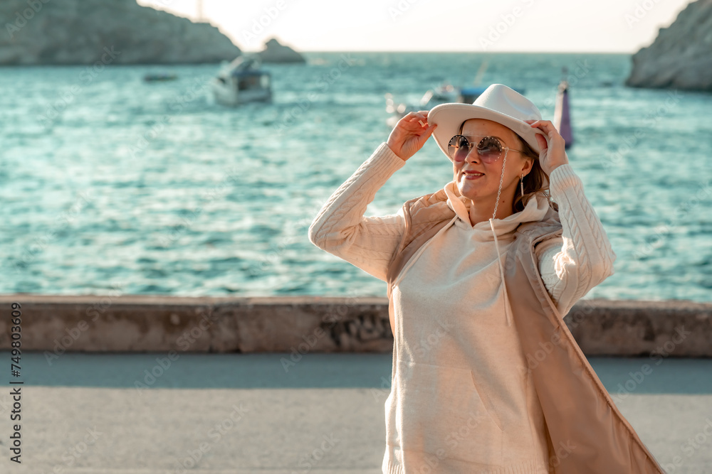 Happy blonde woman in a white suit and hat posing at the camera against the backdrop of the sea