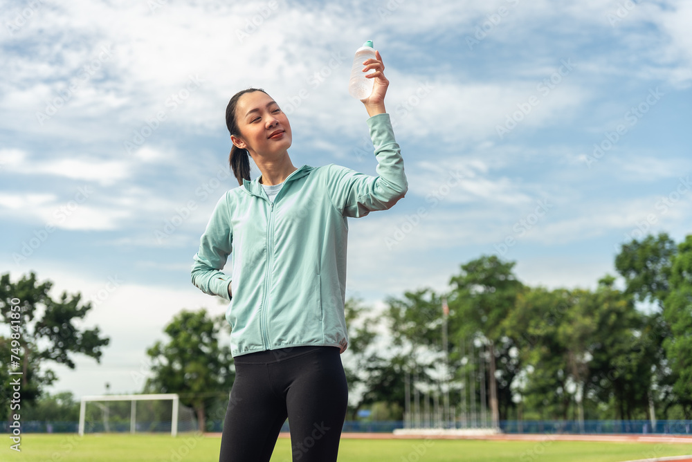 Beautiful asian woman holding drinking water bottle after running, She feeling fresh with sky background, outdoor workout concept, drink clear mineral water after jogging. Generation z lifestyle.	