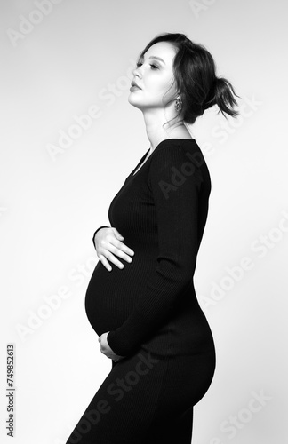 Black and white portrait of pregnant female in black dress with hands near pregnant belly.